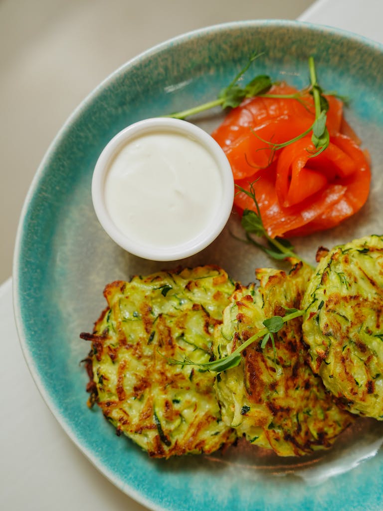 Top view of savory zucchini fritters with sauce and tomato on a ceramic plate.